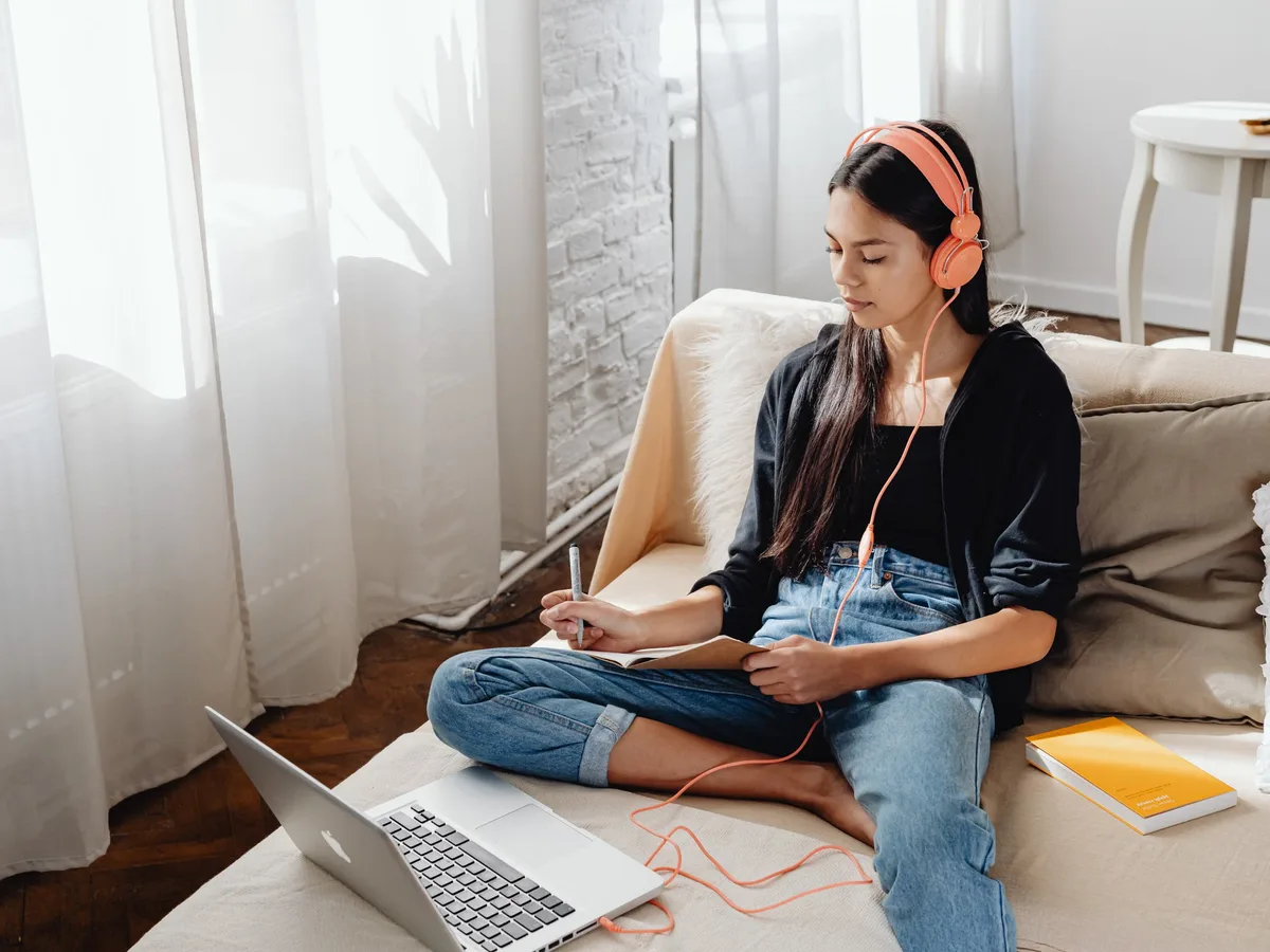 Woman wearing headphones and taking notes beside a laptop